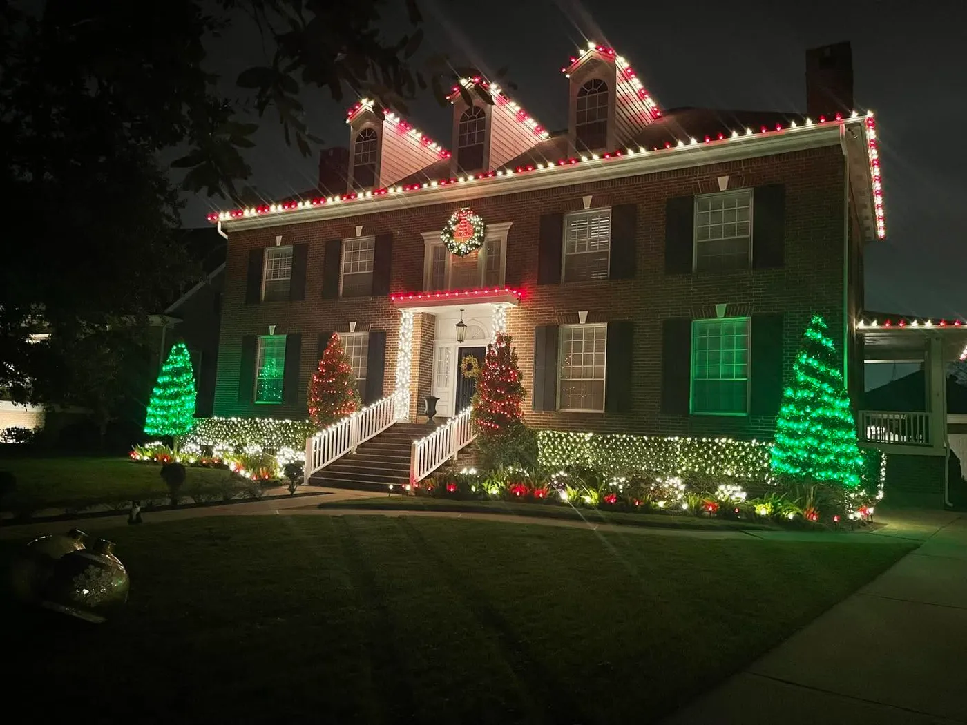 Two-story colonial house with three gables on the roof, wrapped in Christmas lights, landscaping also